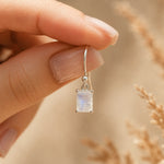 Silver french hook earring with a rectangular emerald cut natural moonstone gemstone held by a hand against a blurred natural background