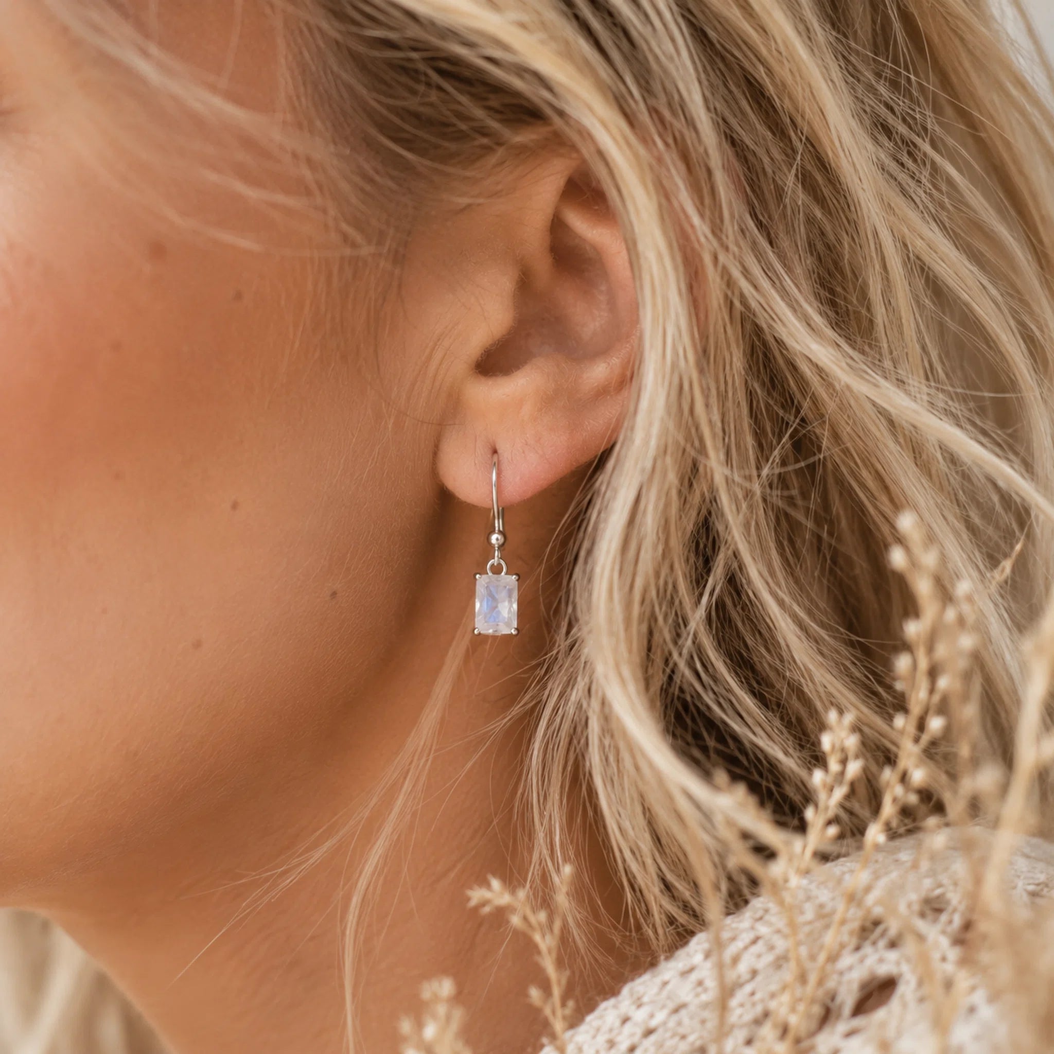 Close-up of a person wearing a silver moonstone earring with a emerald-cut moonstone, surrounded by natural elements.