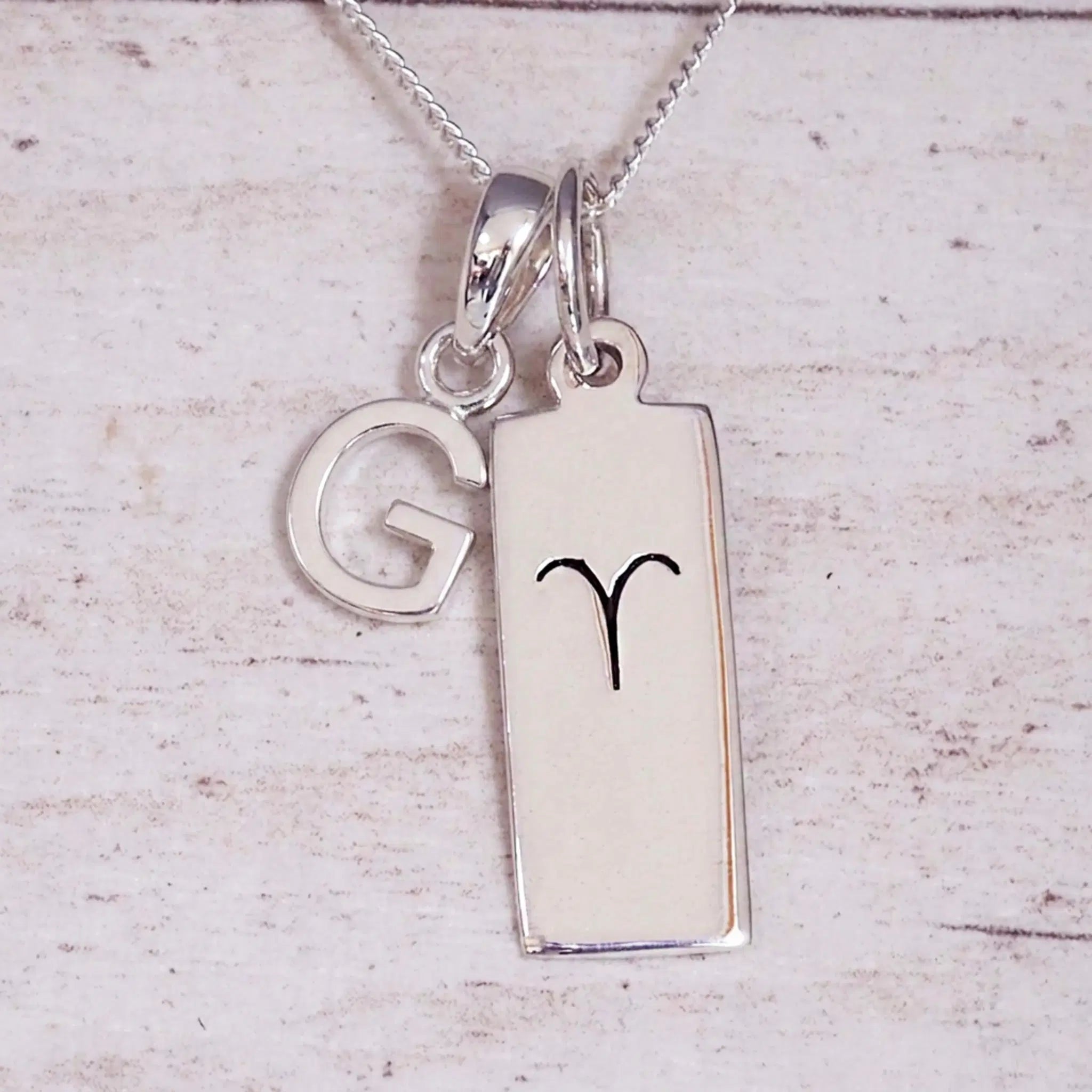 Sterling Silver G Initial pendant and Aries necklace on a fine silver chain on a light wooden background