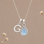 Sterling silver initial G and November natural blue topaz necklace on a taupe background with sparse ivory flowers