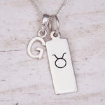 Silver necklace with a pendant featuring the letter 'G' and a Taurus astrological symbol on a light wooden background.
