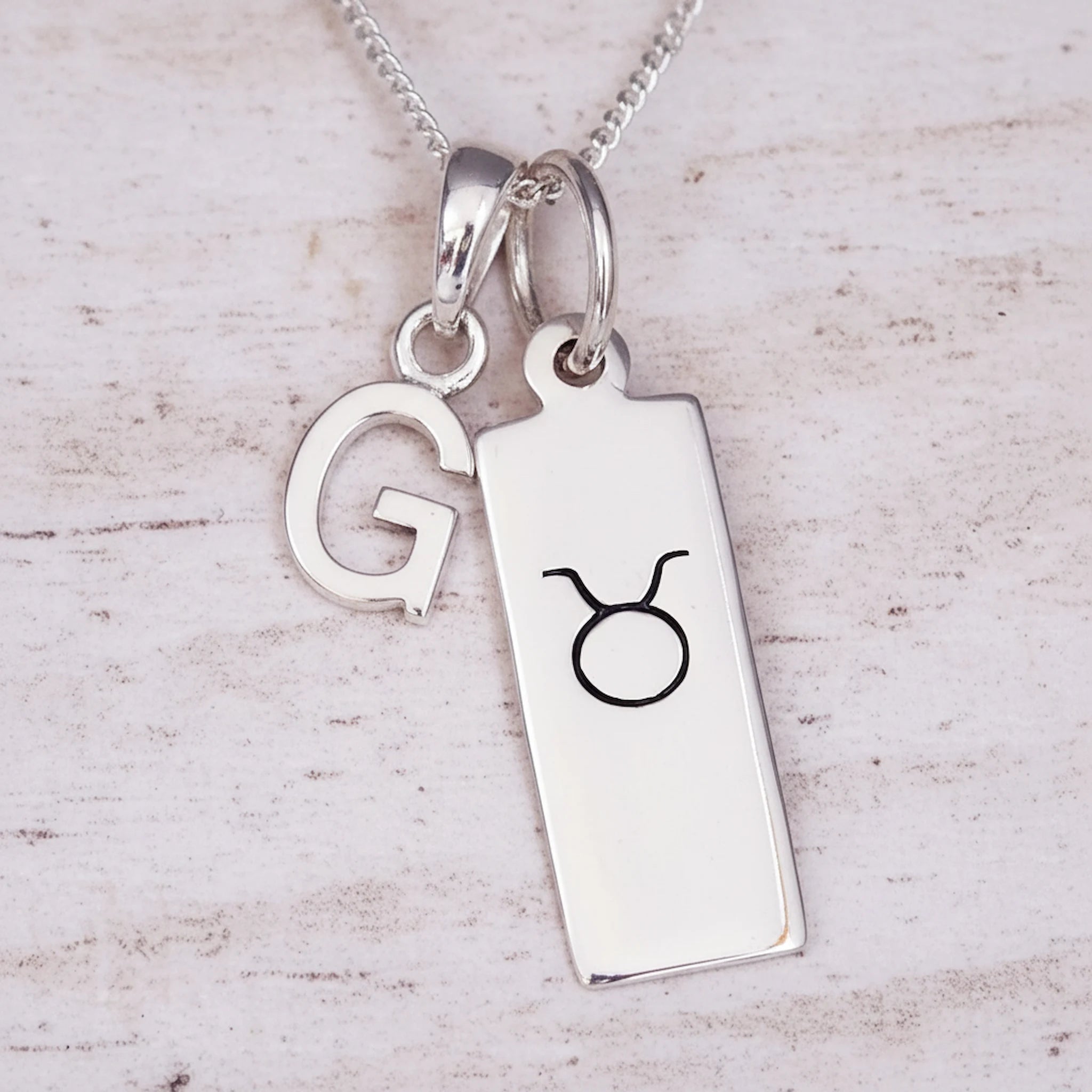 Silver necklace with a pendant featuring the letter 'G' and a Taurus astrological symbol on a light wooden background.