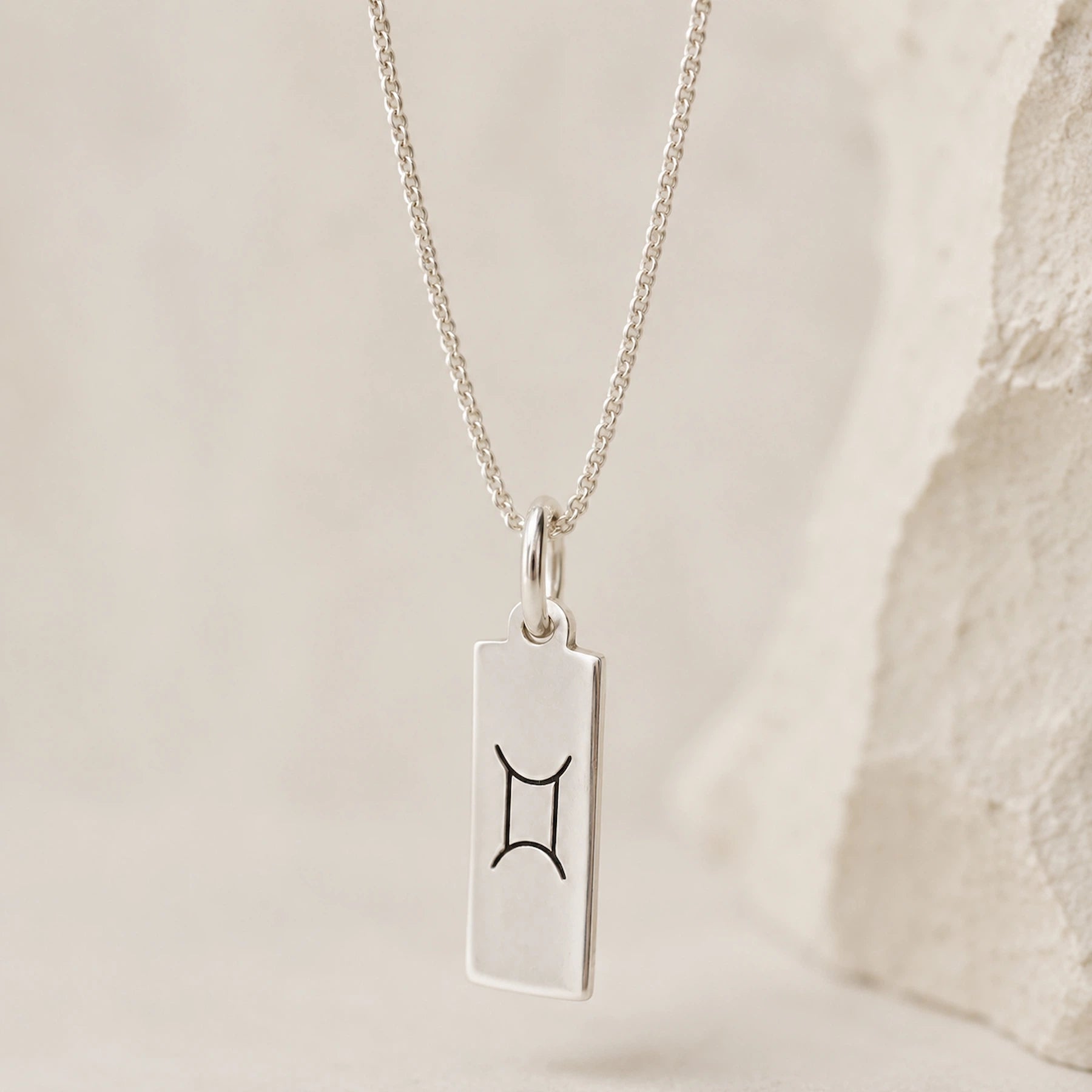 sterling silver fine chain necklace with a rectangle pendant engraved with the gemini zodiac line glyph symbol hanging.
