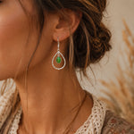 Close-up of a woman wearing a silver earring with a green turquoise gemstone against a blurred natural background.