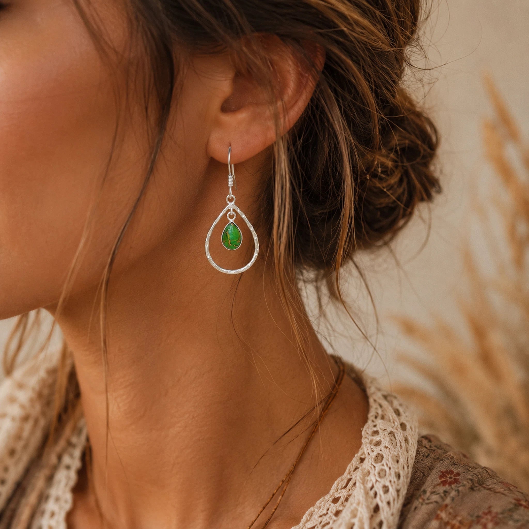 Close-up of a woman wearing a silver earring with a green turquoise gemstone against a blurred natural background.