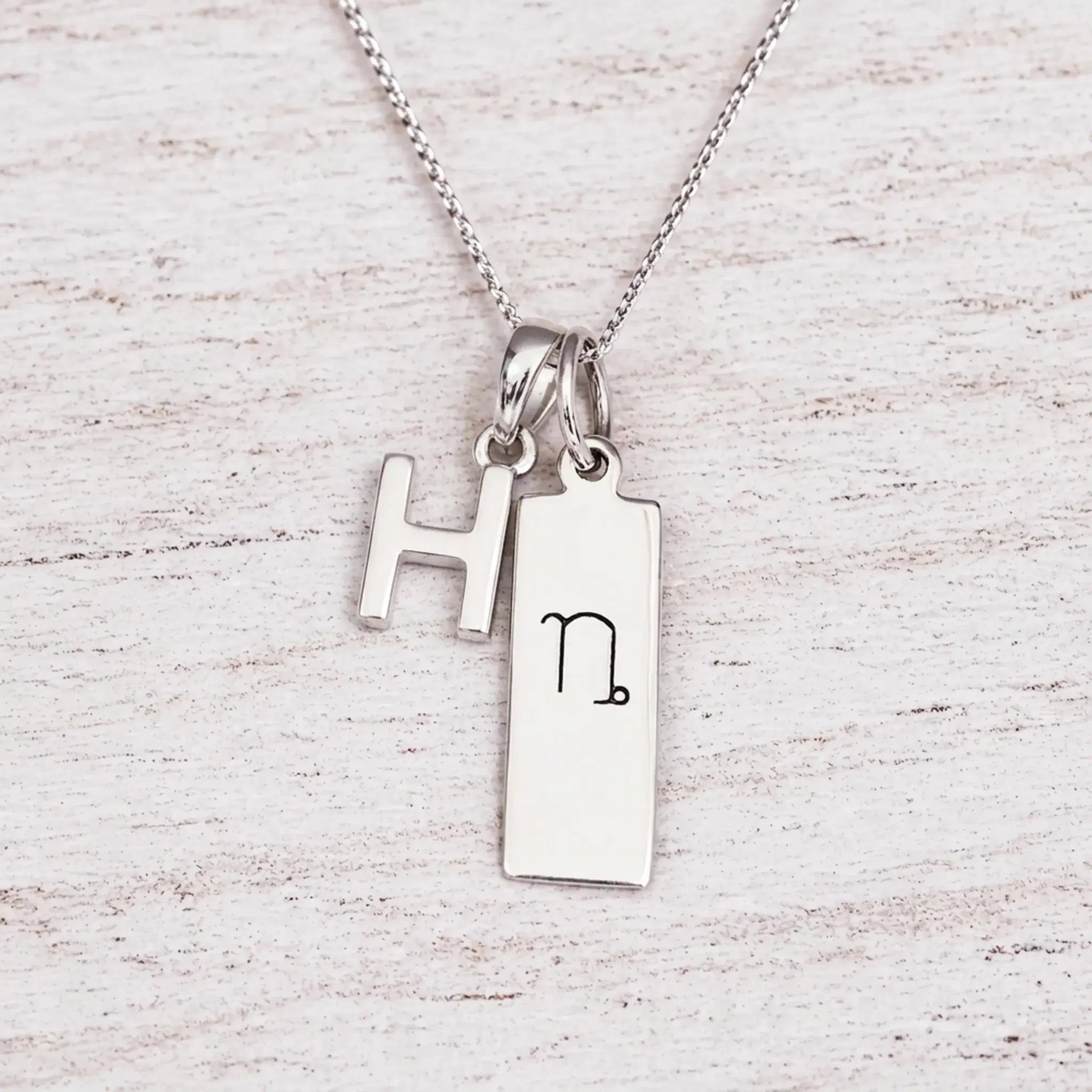 sterling silver initial H pendant and capricorn necklace on a fine silver chain against a light wooden background