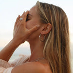 Woman with hand on face against a blurred natural background wearing large stainless steel silver hoop earrings