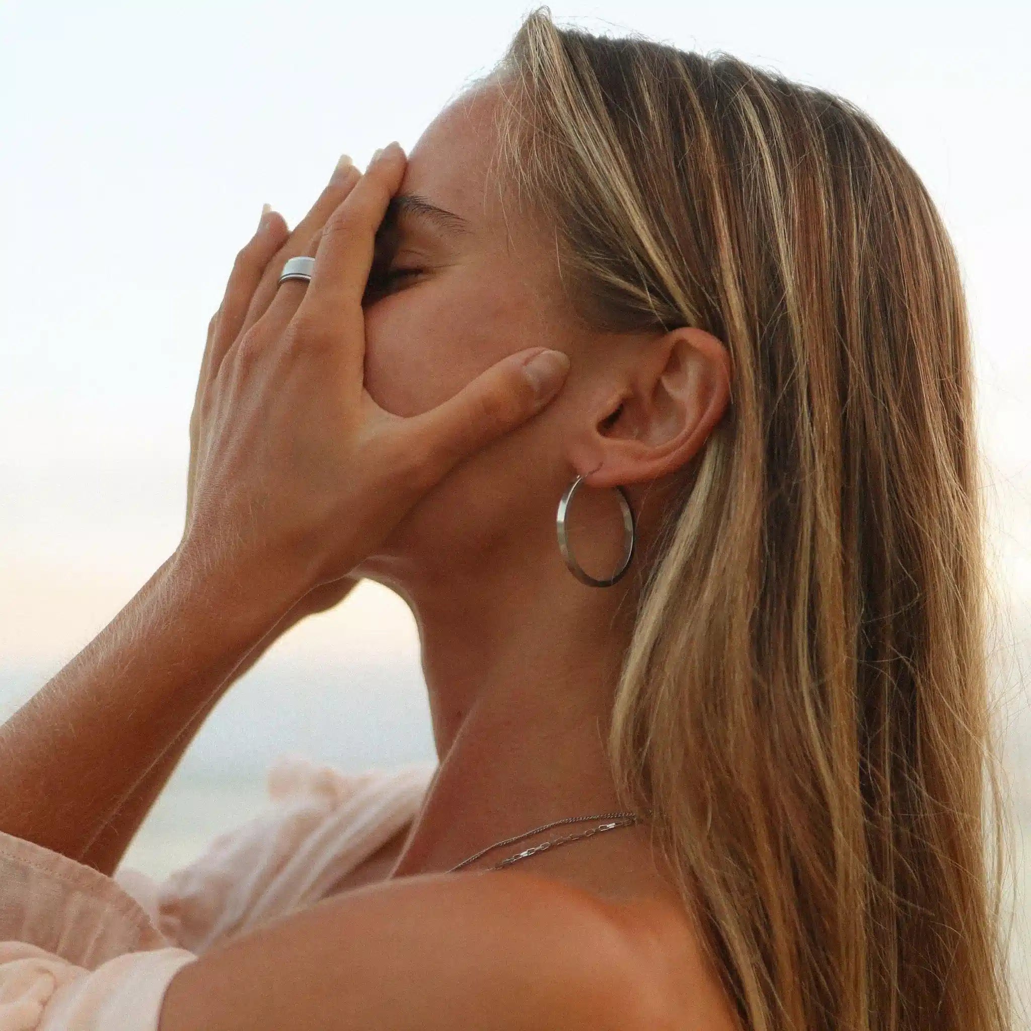 Woman with hand on face against a blurred natural background wearing large stainless steel silver hoop earrings