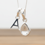 Silver necklace with 'A' initial pendant and teardrop-shaped Herkimer Quartz Crystal hanging above a wooden table