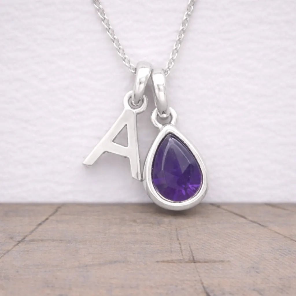 Silver necklace with a teardrop-shaped purple amethyst gemstone and letter 'A' pendant on a light background.