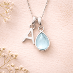 sterling silver Necklace with a teardrop-shaped february birthstone aqumarine pendant and letter 'A' charm on a soft pink background with delicate flowers.