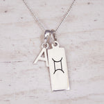 Sterling Silver double pendant necklace with a rectangular gemini zodiac glyph pendant and a upper case block letter 'A' pendant on a light wooden background.