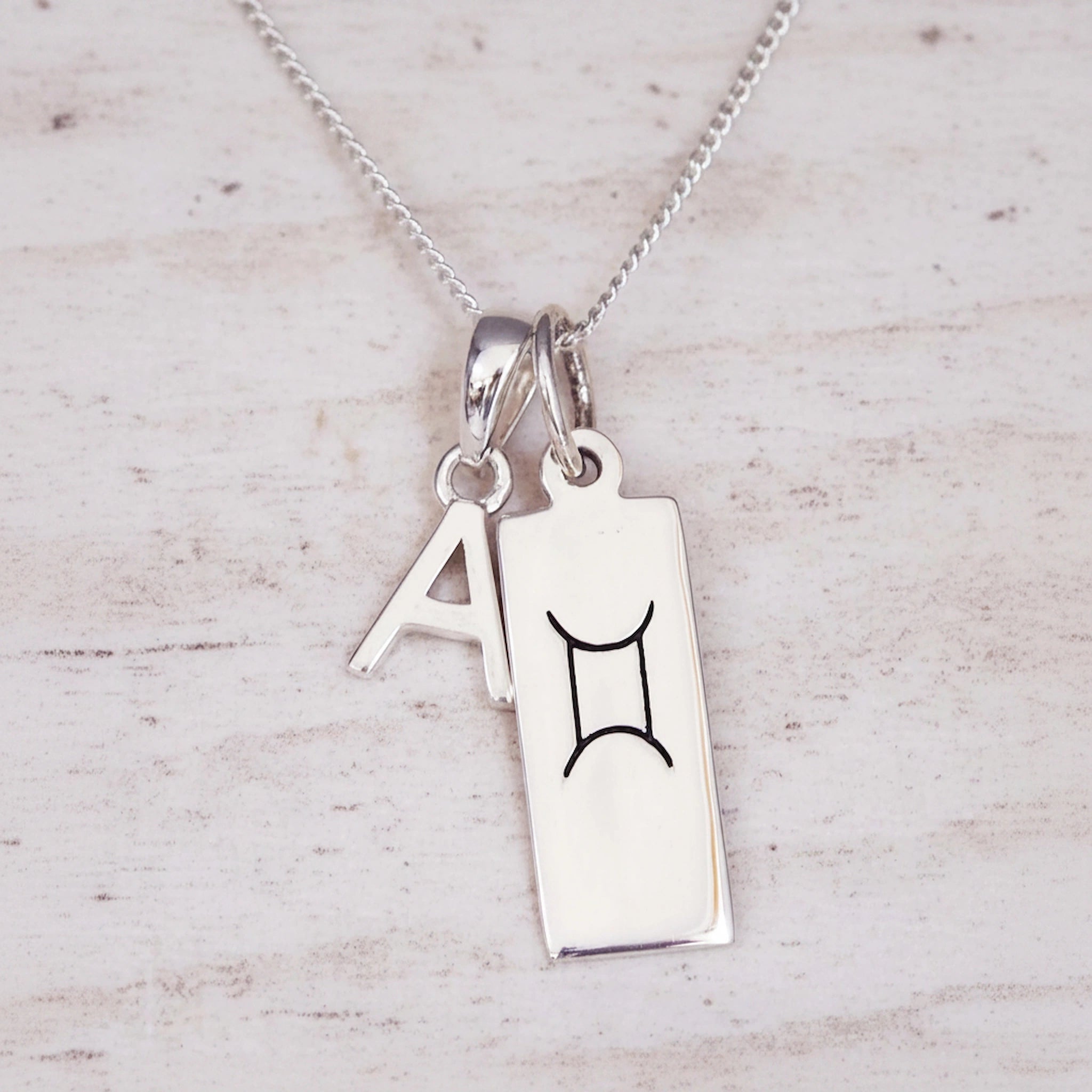 Sterling Silver double pendant necklace with a rectangular gemini zodiac glyph pendant and a upper case block letter 'A' pendant on a light wooden background.