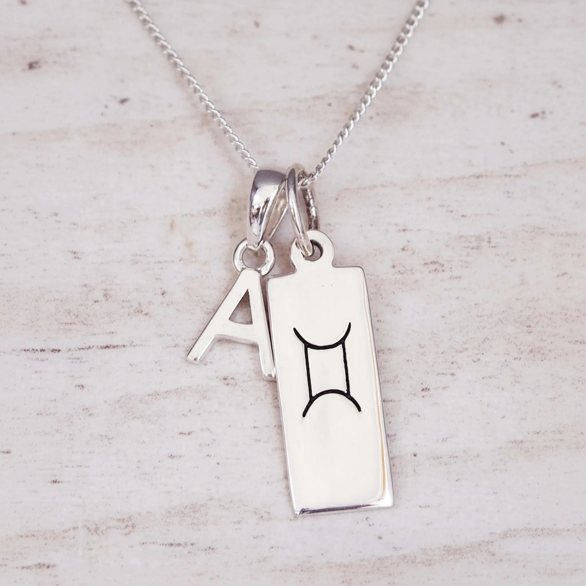 Sterling Silver double pendant necklace with a rectangular gemini zodiac glyph pendant and a upper case block letter 'A' pendant on a light wooden background.