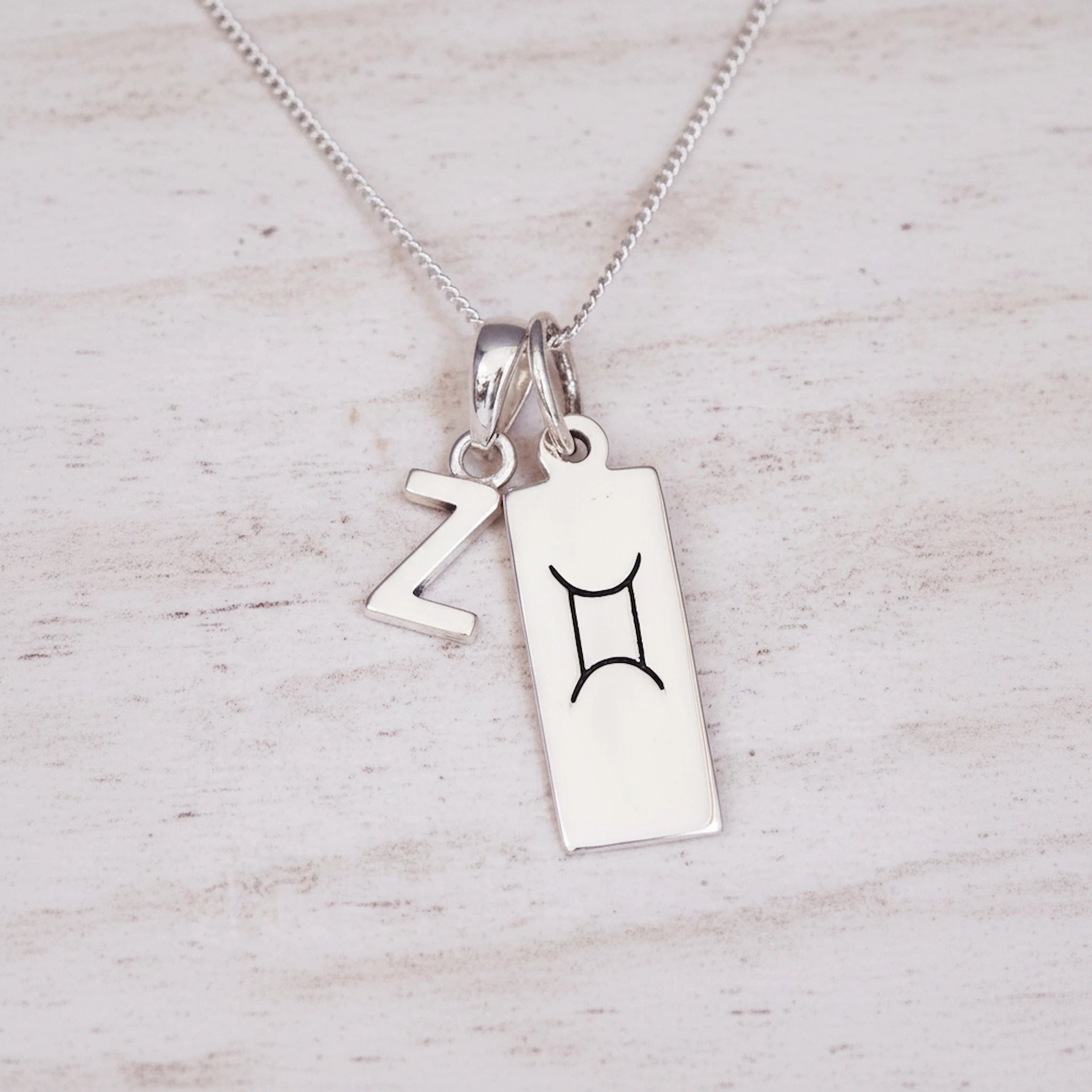 Sterling Silver double pendant necklace with a rectangular gemini zodiac glyph pendant and a upper case block letter 'Z' pendant on a light wooden background.