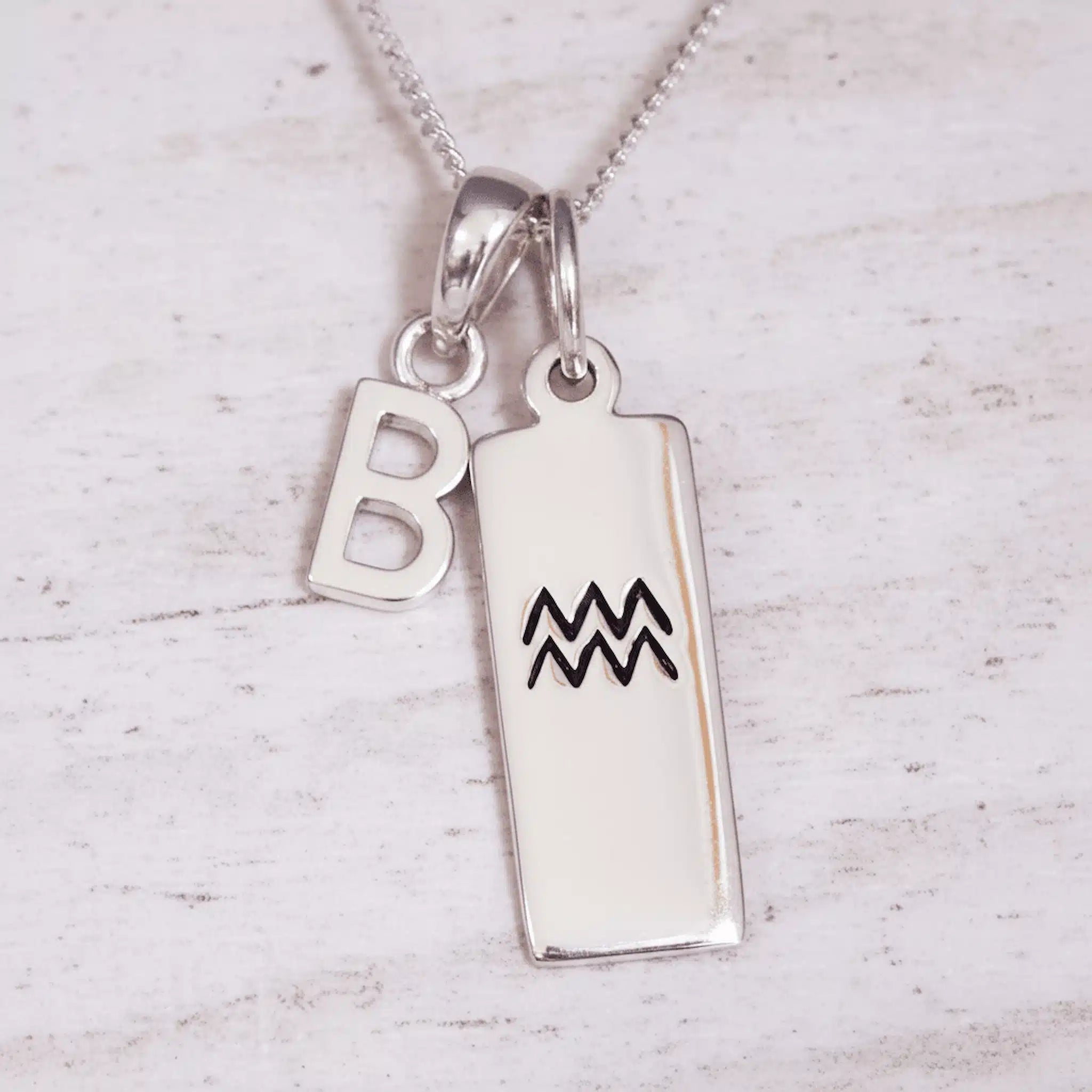 Silver necklace with 'B' pendant and Aquarius zodiac symbol pendant on a light wooden background