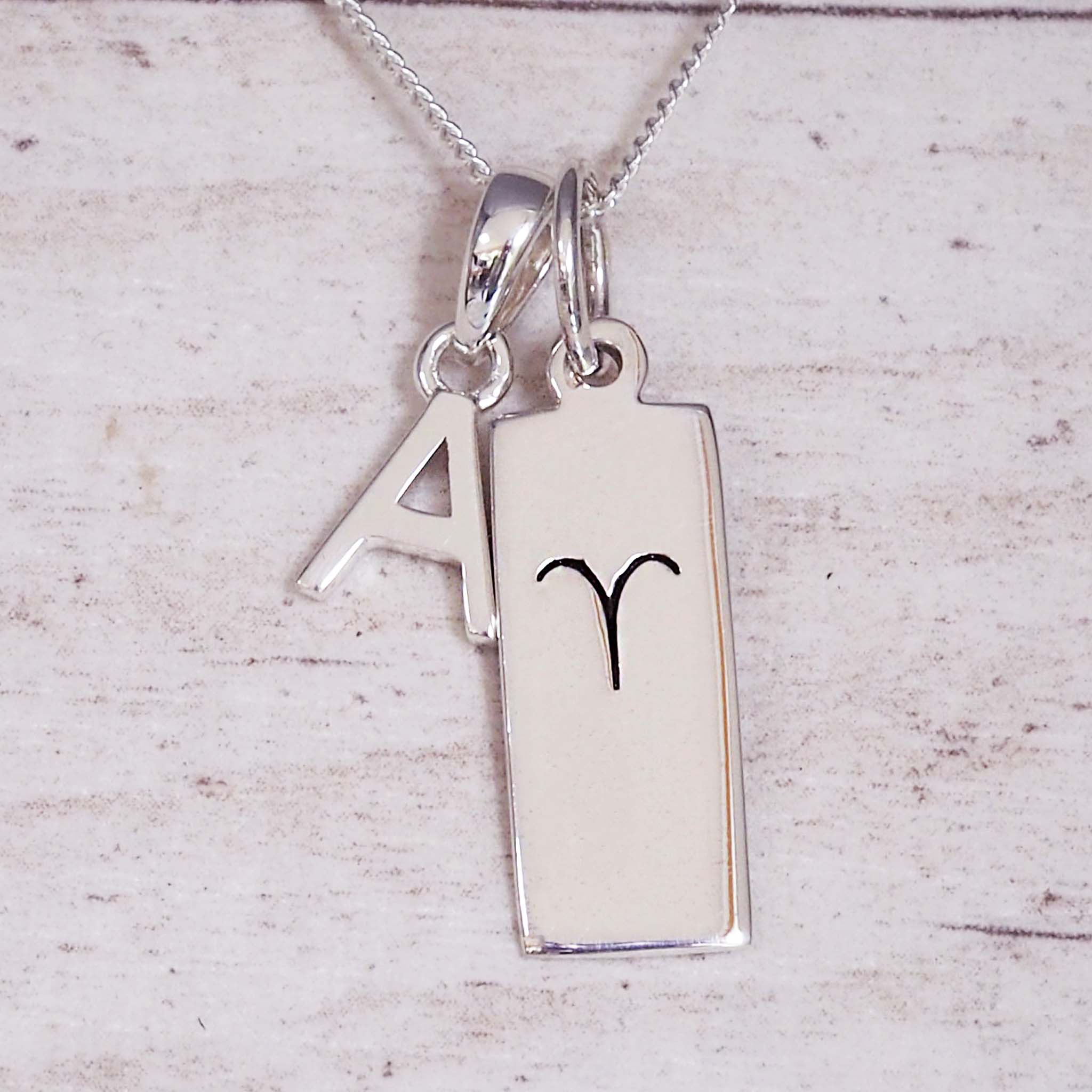 Silver necklace with a ‘A’ initial pendant and a rectangular Aries zodiac pendant on a light wooden background