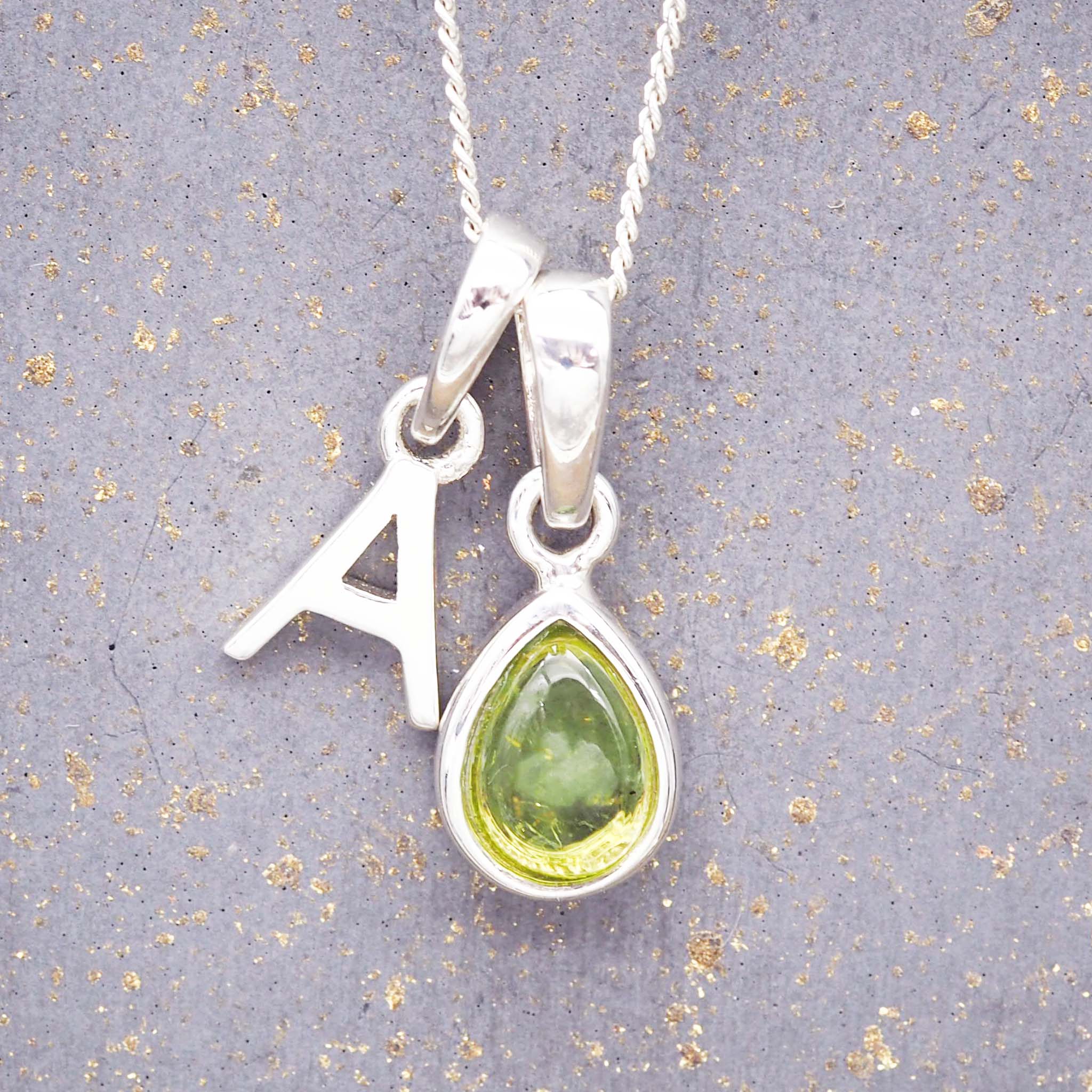 Silver necklace with a 'A' initial pendant and a green teardrop august birthstone peridot pendant on a grey background