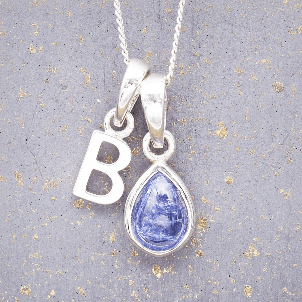 Silver necklace with a teardrop-shaped december birthstone tanzanite pendant and 'B' charm on a textured gray background.