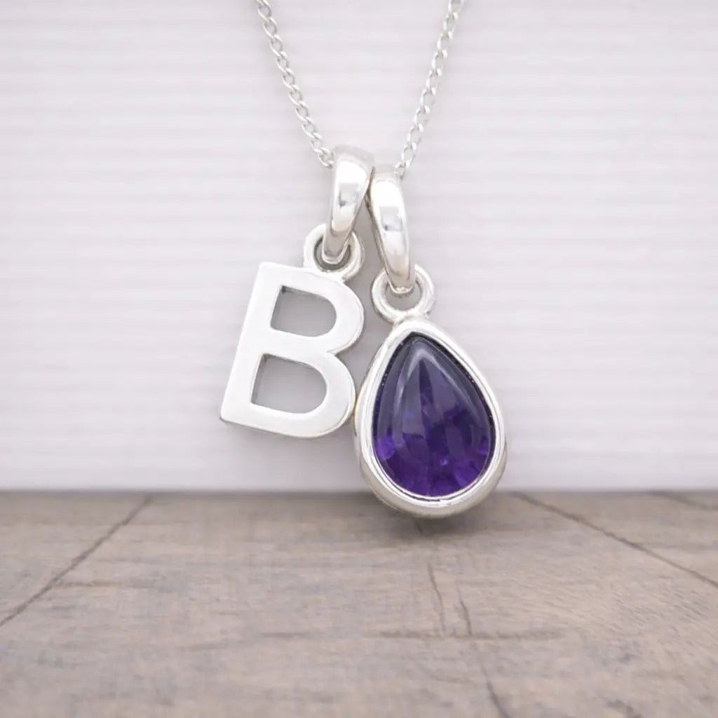 Silver necklace with 'B' pendant and purple teardrop-shaped amethyst gemstone on a wooden surface.