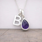 Silver necklace with 'B' pendant and purple teardrop-shaped amethyst gemstone on a wooden surface.