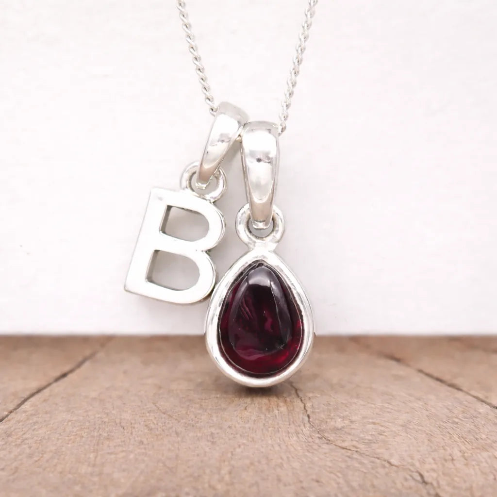 Silver necklace with a teardrop-shaped red garnet gemstone pendant and a letter 'B' charm on a light background.