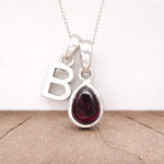 Silver necklace with a teardrop-shaped red garnet gemstone pendant and a letter 'B' charm on a light background.