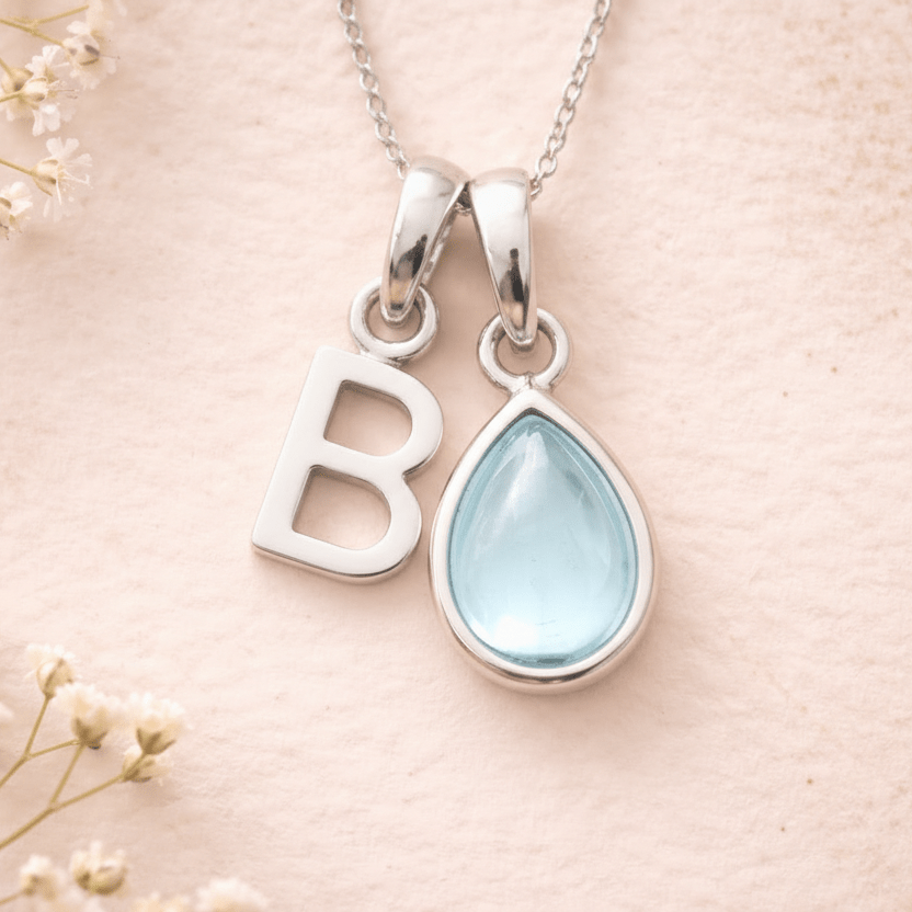 Silver necklace with 'B' pendant and teardrop-shaped light blue march birthstone aquamarine gemstone on a soft pink background.