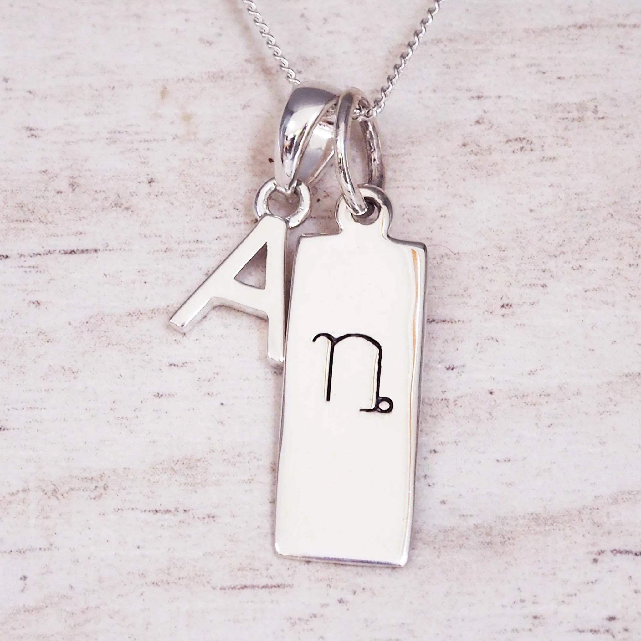 sterling Silver double pendant necklace with a rectangle pendant with the capricorn zodiac glyph, and a 'A' initial pendant on a light wooden background