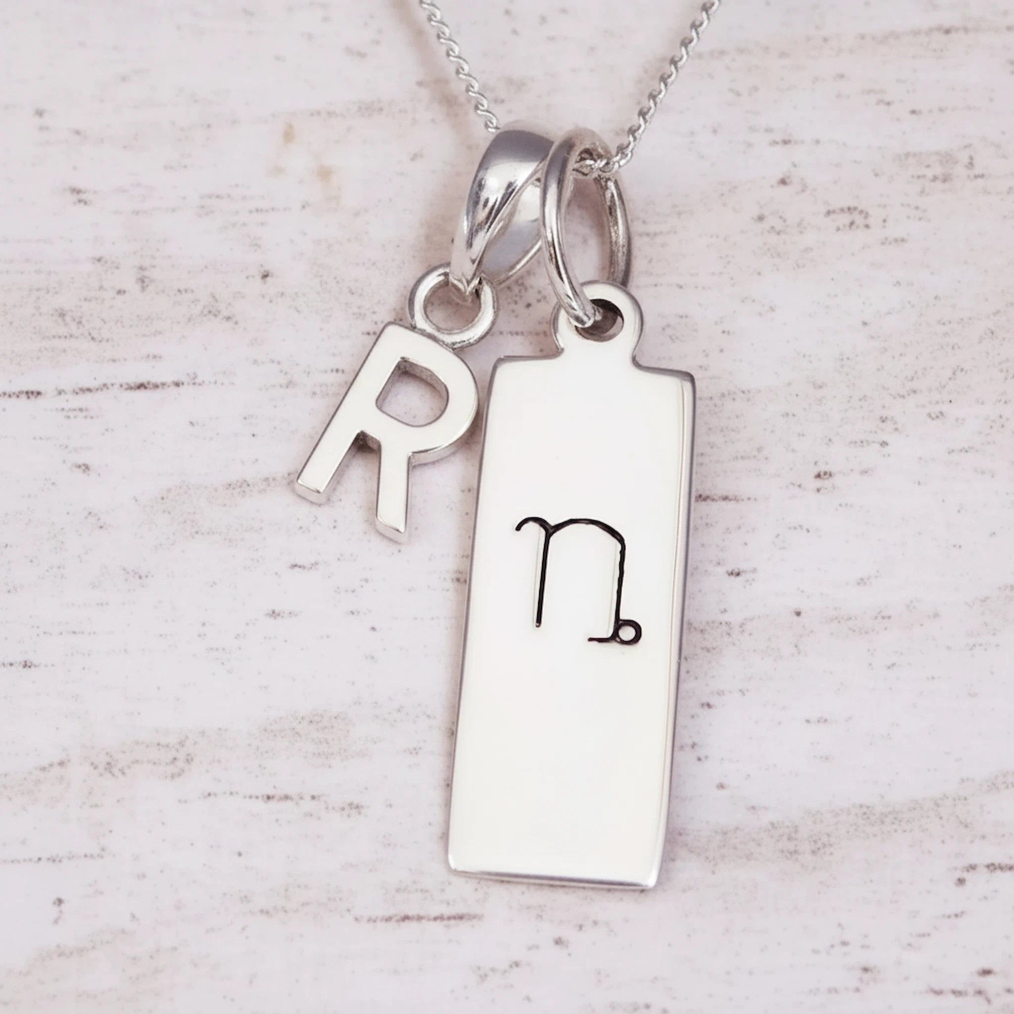 sterling Silver double pendant necklace with a rectangle pendant with the capricorn zodiac glyph, and a 'R' initial pendant on a light wooden background