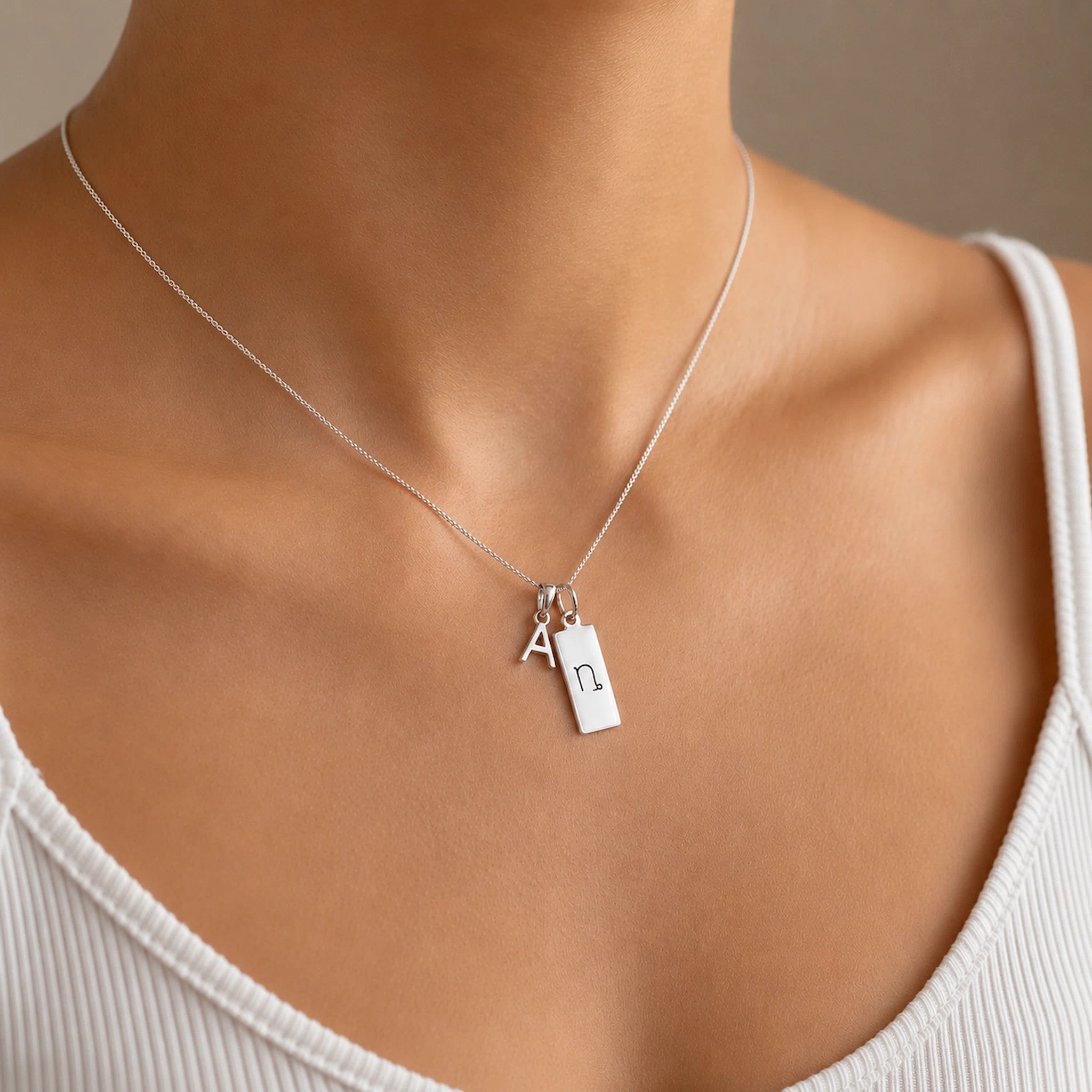 model wearing a sterling Silver double pendant necklace with a rectangle pendant with the capricorn zodiac glyph, and a initial pendant on a light wooden background