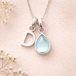 Silver necklace with 'D' pendant and teardrop-shaped light blue stone on a pink background with small white flowers.