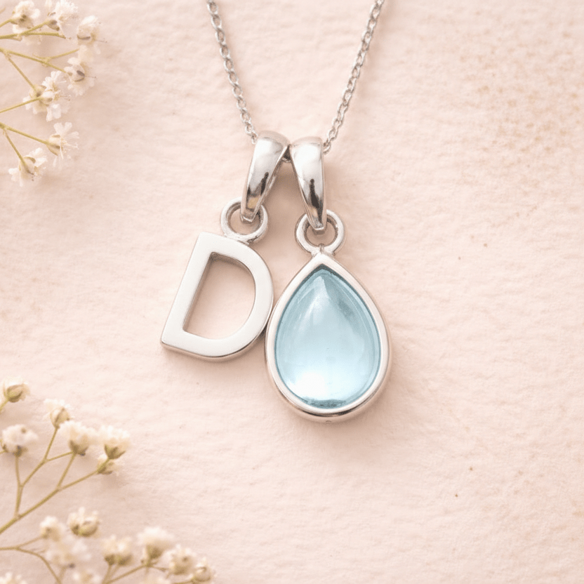 Silver necklace with 'D' pendant and teardrop-shaped light blue stone on a pink background with small white flowers.
