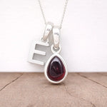 Silver Initial E necklace with a teardrop-shaped Garnet gemstone pendant hanging above a rustic wooden surface
