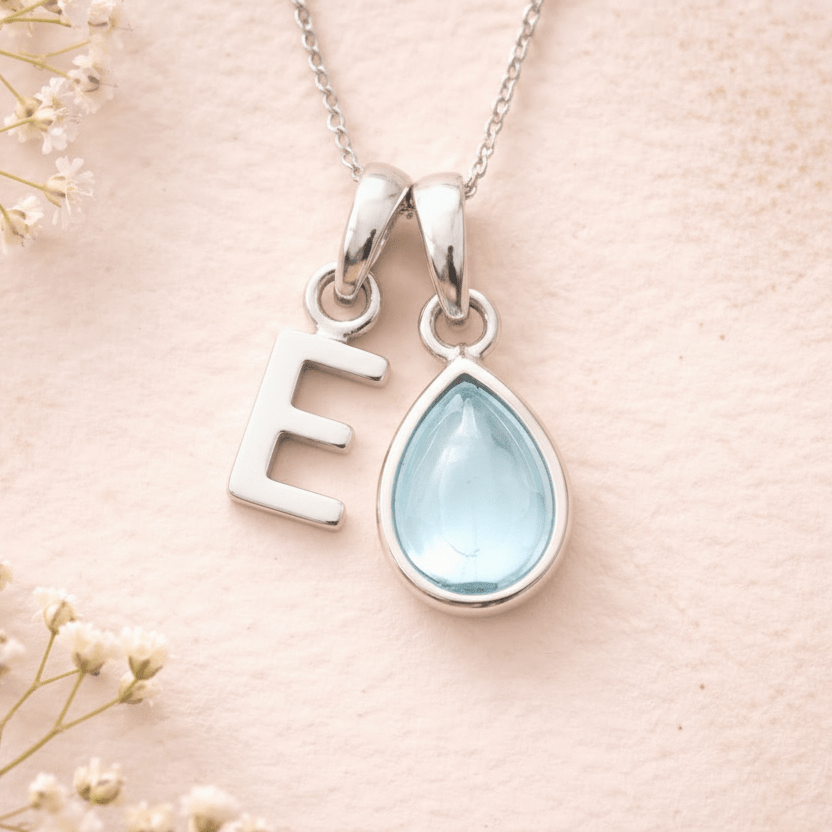 sterling silver Necklace with a teardrop-shaped february birthstone aqumarine pendant and letter 'E' charm on a soft pink background with delicate flowers.