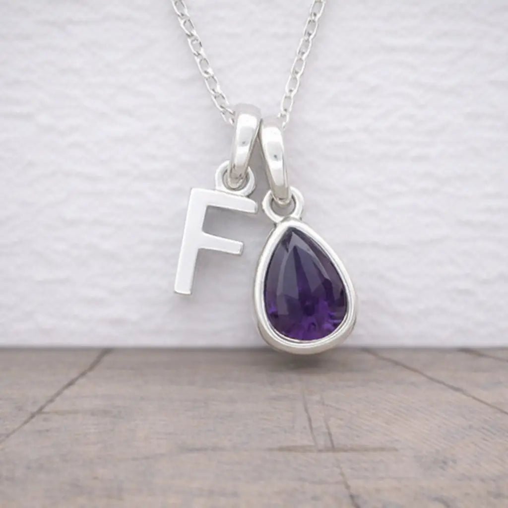 Silver necklace with a teardrop-shaped purple amethyst gemstone and letter 'F' pendant on a light background.