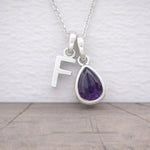 Silver necklace with a teardrop-shaped purple amethyst gemstone and letter 'F' pendant on a light background.