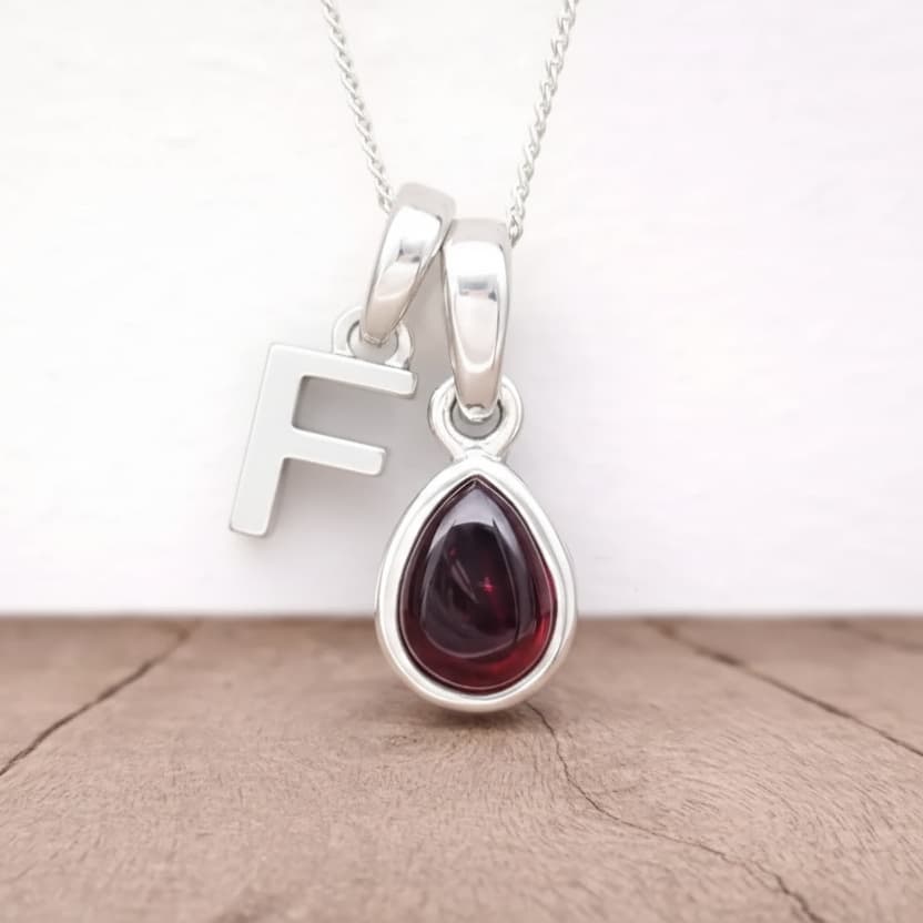 Silver initial F necklace with a teardrop-shaped red garnet gemstone pendant on a beige surface.