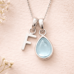 sterling silver Necklace with a teardrop-shaped aquamarine pendant and letter 'F' charm on a soft pink background.