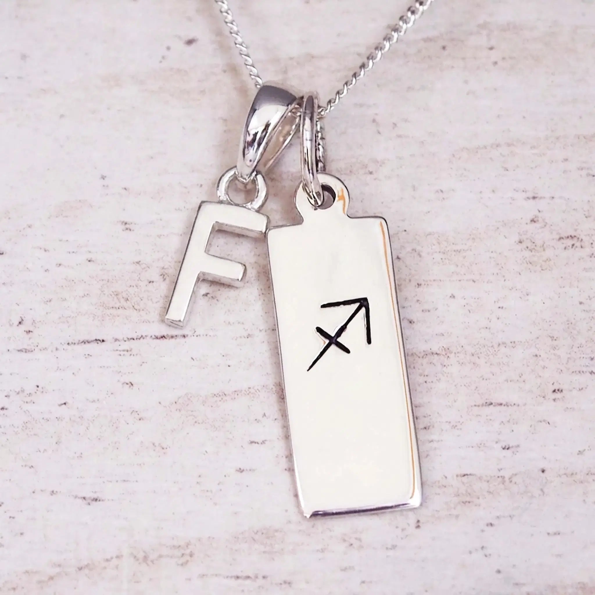 Sterling silver initial F and Sagittarius zodiac bar necklace, close-up of the letter charm and pendant on whitewashed wood