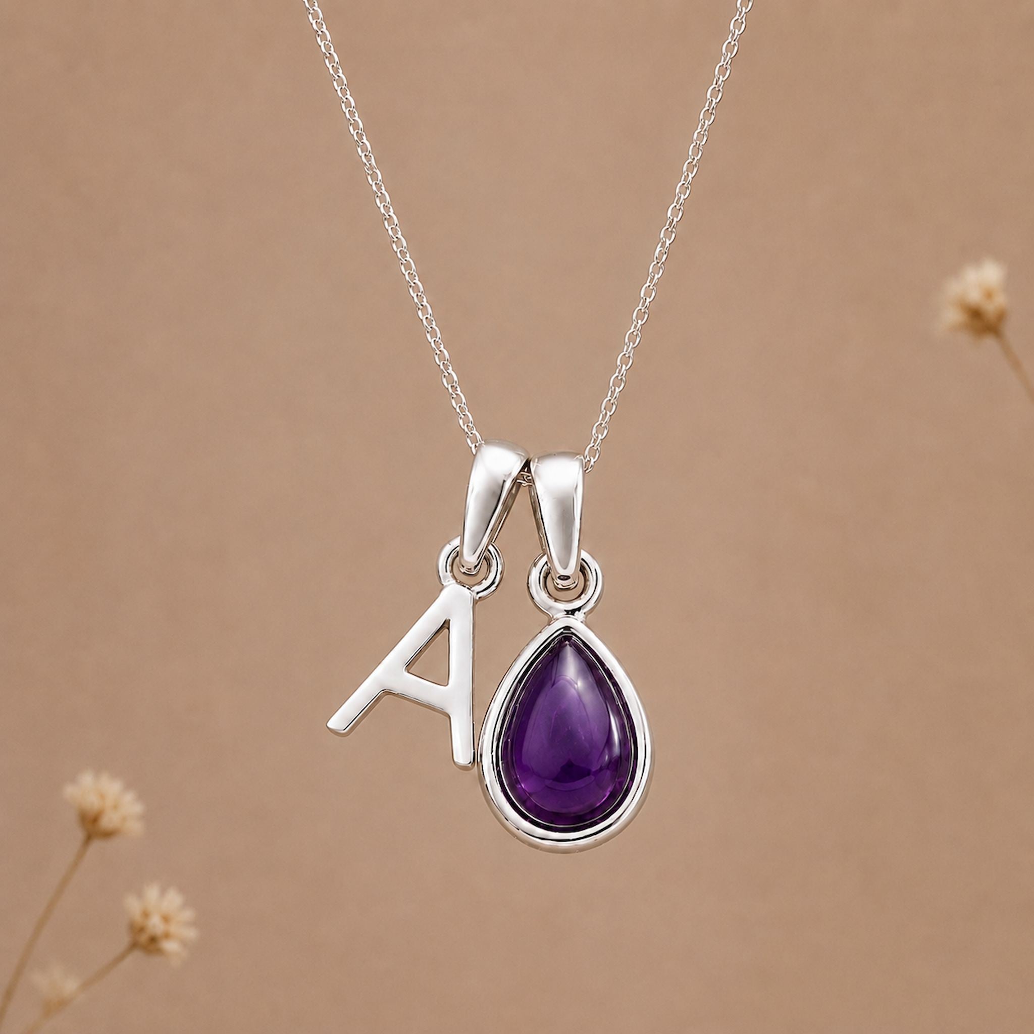A silver initial pendant necklace featuring the letter 'A' next to a teardrop-shaped purple amethyst gemstone pendant, shown against a warm brown background with subtle dried flowers.