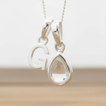 Silver Initial G necklace with a teardrop-shaped Herkimer Quartz crystal pendant hanging above a rustic wooden surface