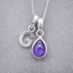 sterling Silver necklace with a 'G' initial pendant and a teardrop-shaped purple february birthstone amethyst gemstone on a grey background.