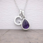 Silver necklace with 'G' pendant and teardrop-shaped purple amethyst gemstone on a light background