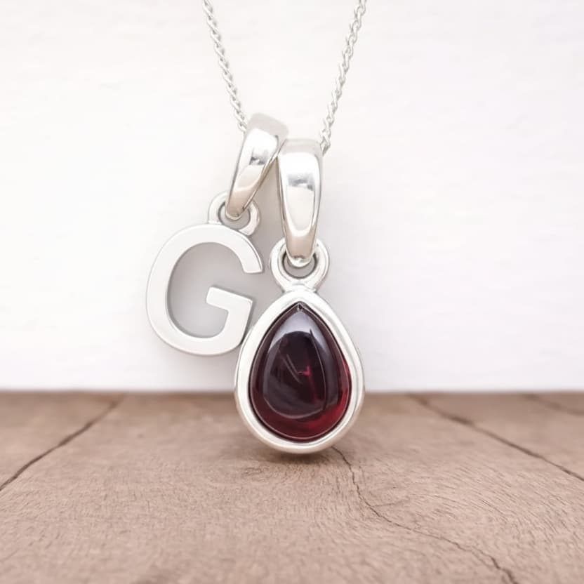 Silver initial G necklace with a teardrop-shaped garnet gemstone pendant on a light wooden surface.