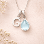 Necklace with 'G' pendant and teardrop-shaped light blue stone on a soft pink background with delicate flowers.