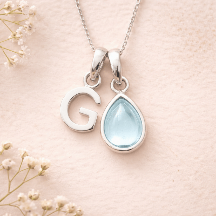 Necklace with 'G' pendant and teardrop-shaped light blue stone on a soft pink background with delicate flowers.