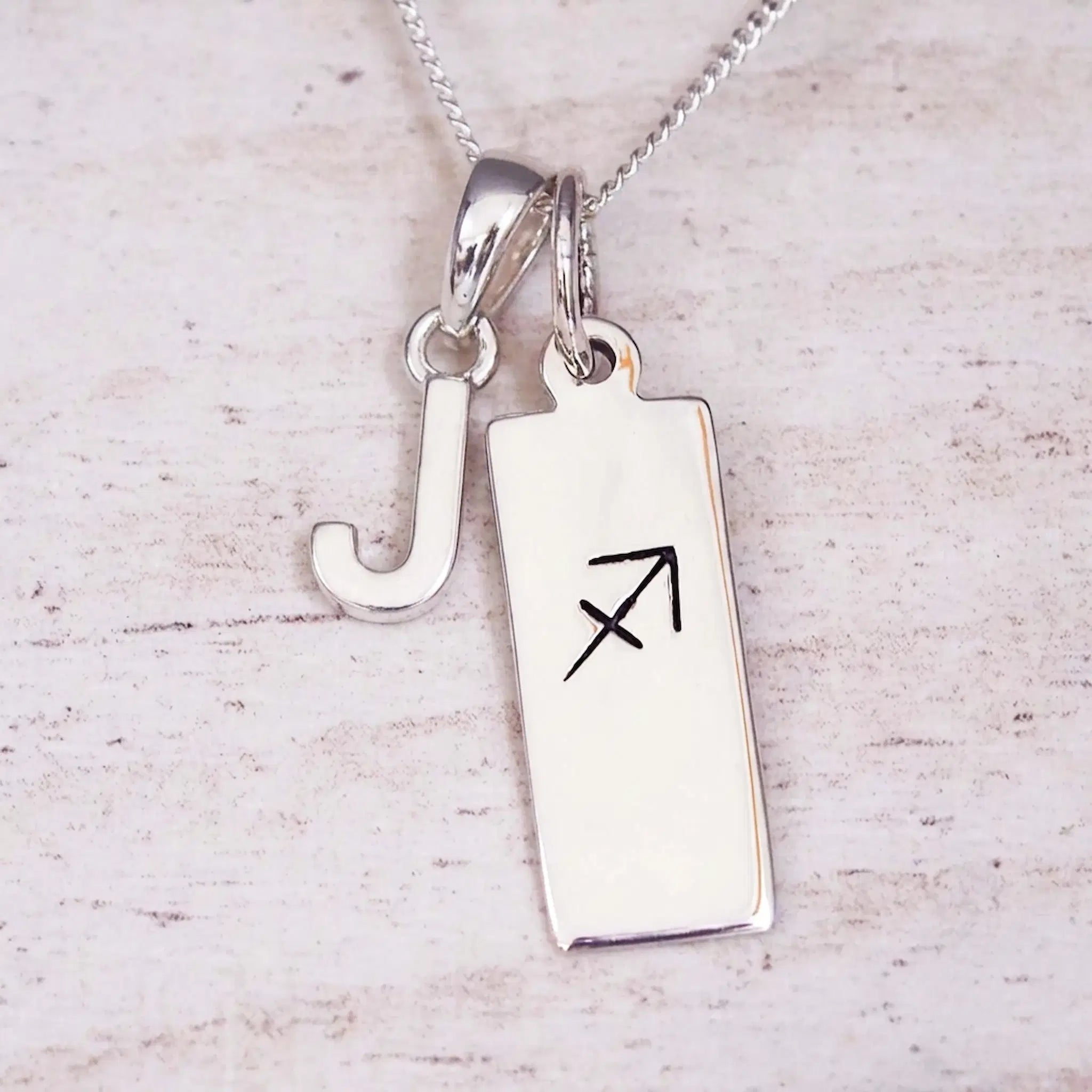 Sterling silver initial J and Sagittarius zodiac bar necklace, close-up of the letter charm and pendant on whitewashed wood