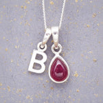 Sterling Silver necklace with 'B' initial pendant and July Birthstone Ruby gemstone pear-shaped pendant on a grey background.