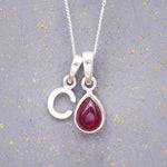 Sterling Silver necklace with 'C' initial pendant and July Birthstone Ruby gemstone pear-shaped pendant on a grey background.